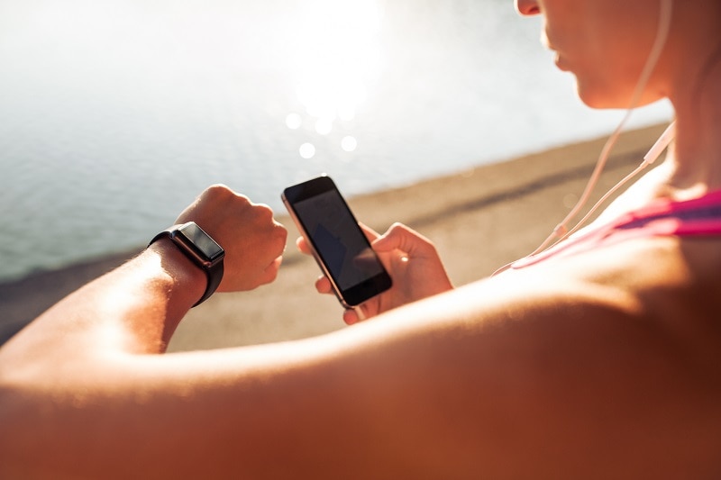 Sportswoman looking at smartwatch and holding smart phone in her other hand, outdoors. Fitness female setting up her smartwatch for her run.