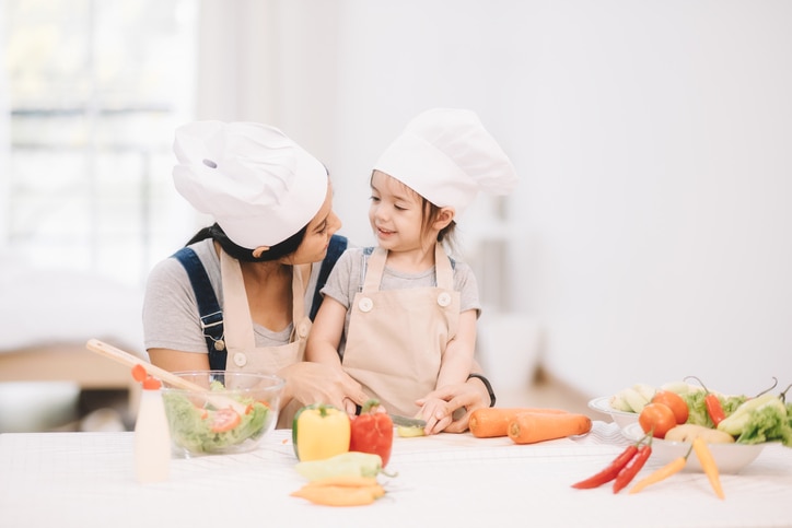 Cute little girl and her mom in chef's hats are cutting vegetables cooking a salad and smiling