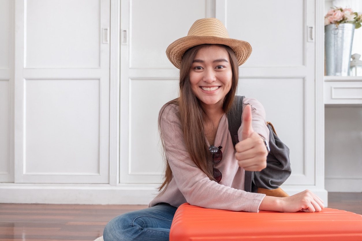 Young asian woman traveler thumbs up with orange suitcase prepare for holiday vacation at home.backpacker travel concept